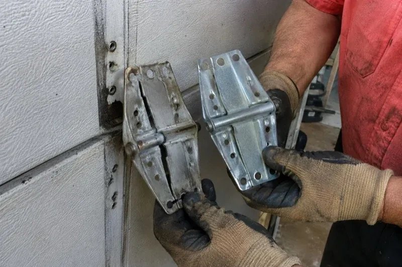 Gloved hands holding two metal garage-door hinges side by side, one discolored with a visible crack and the other bright silver. — Garage Door Hinge Replacement, Strathroy-Caradoc