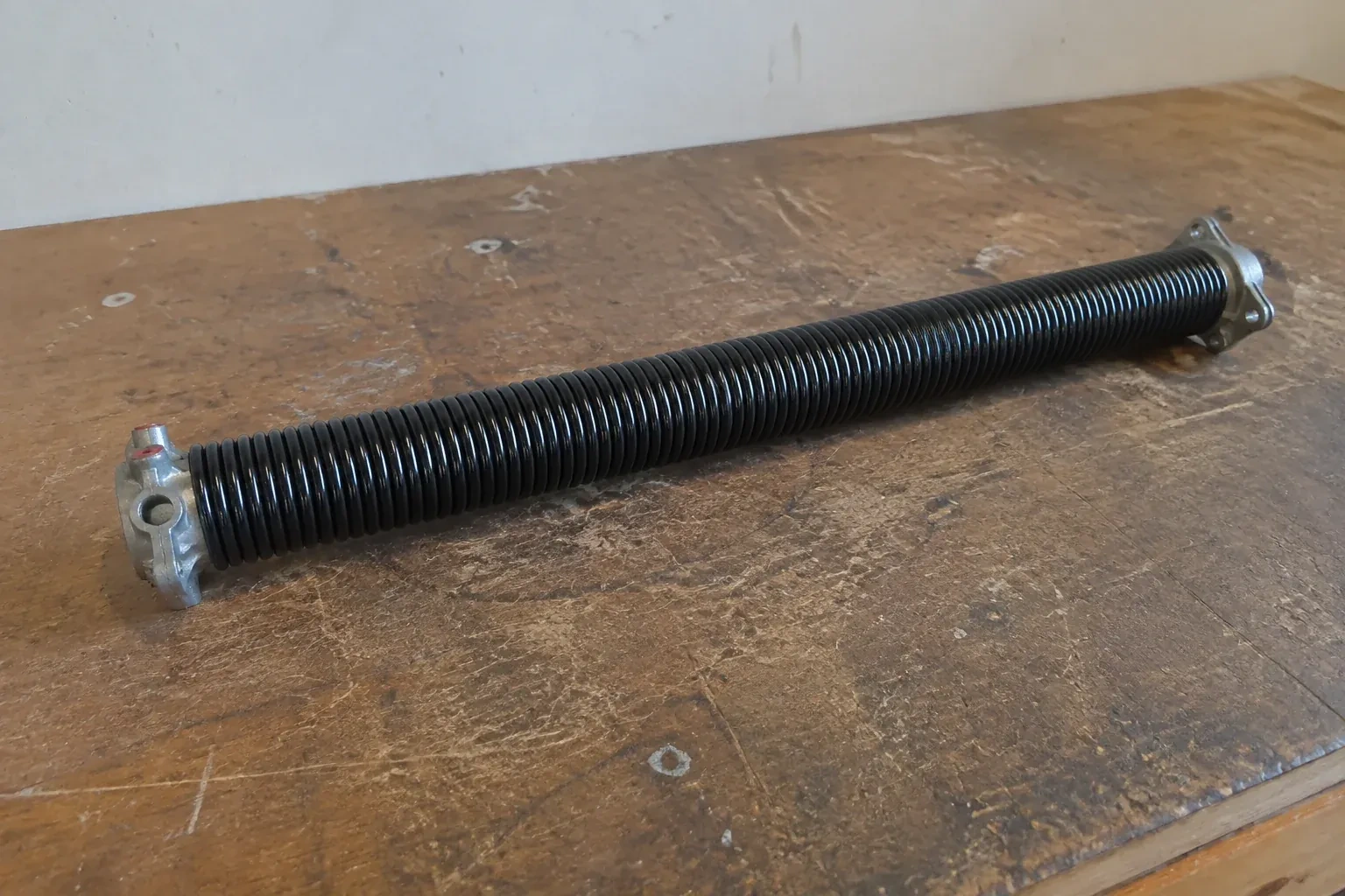 Detached black coil spring with metal end fittings on a workbench. — Torsion Spring Replacement, Strathroy-Caradoc